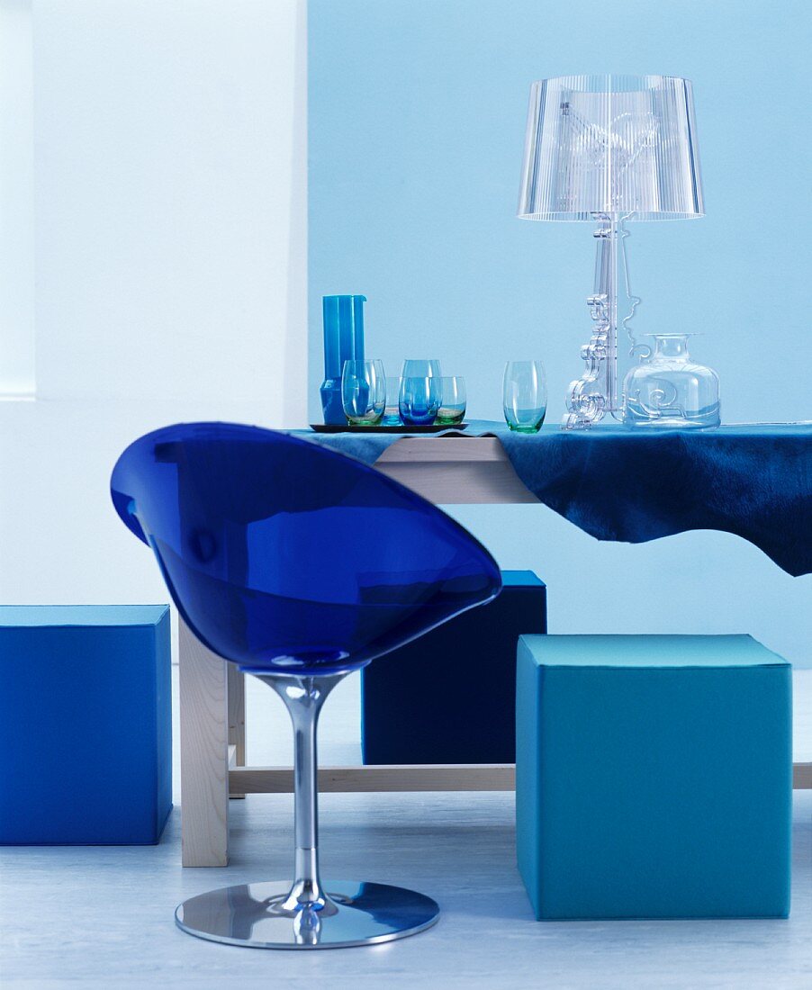 Blue plastic chair and cubic pouffe around glasses and lamp on table