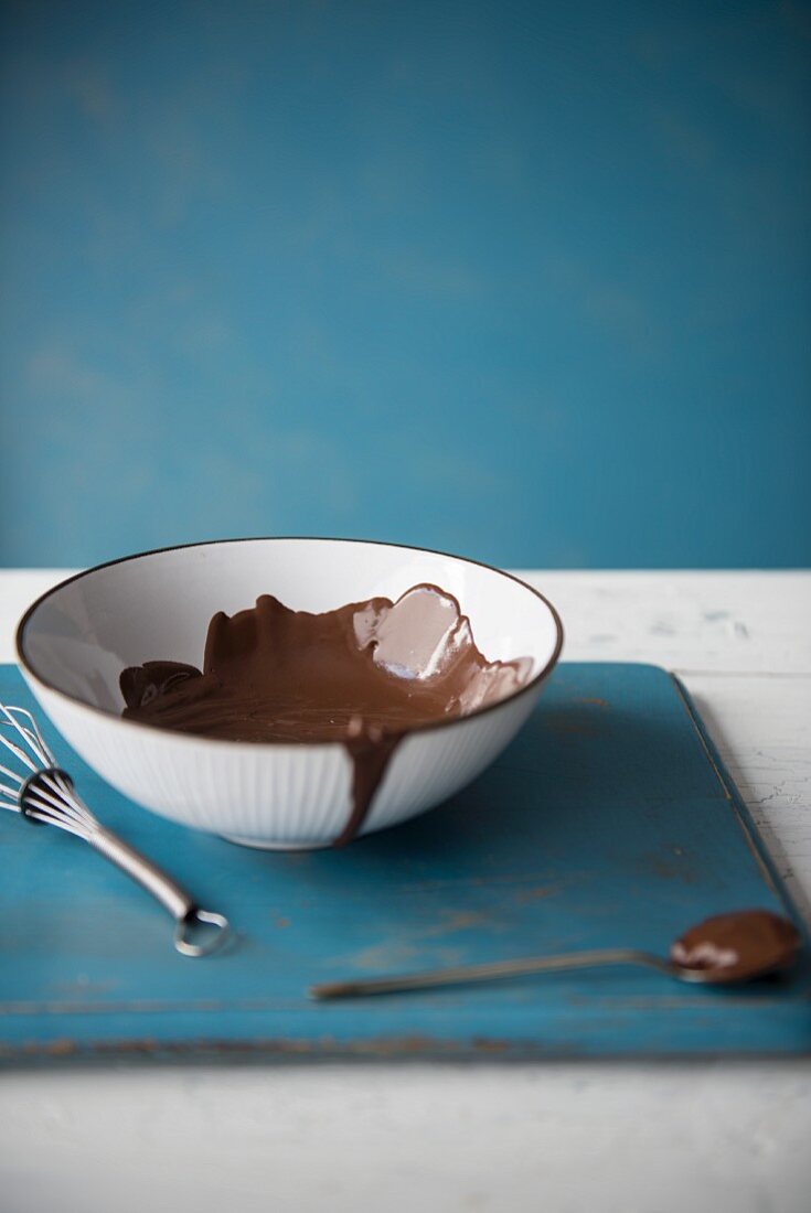 A bowl of chocolate sauce next to a spoon and a whisk
