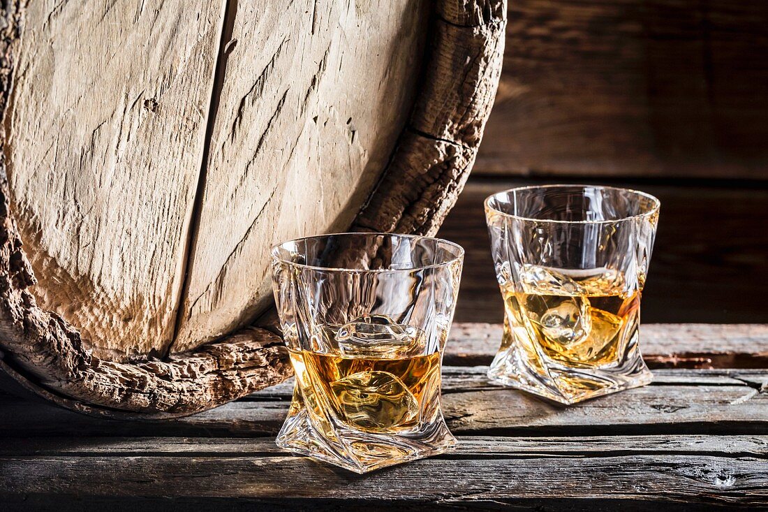 Zwei Gläser Whisky mit Eiswürfeln vor altem Holzfass