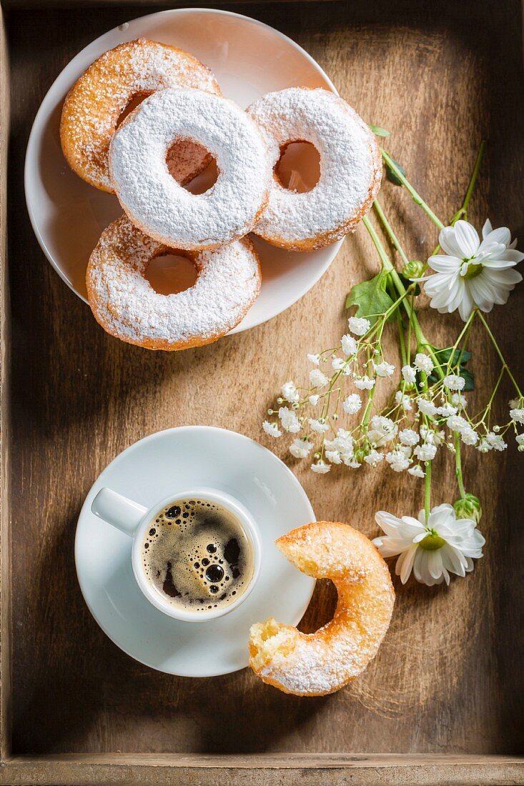Espresso, doughnuts with icing sugar and flowers on a wooden tray