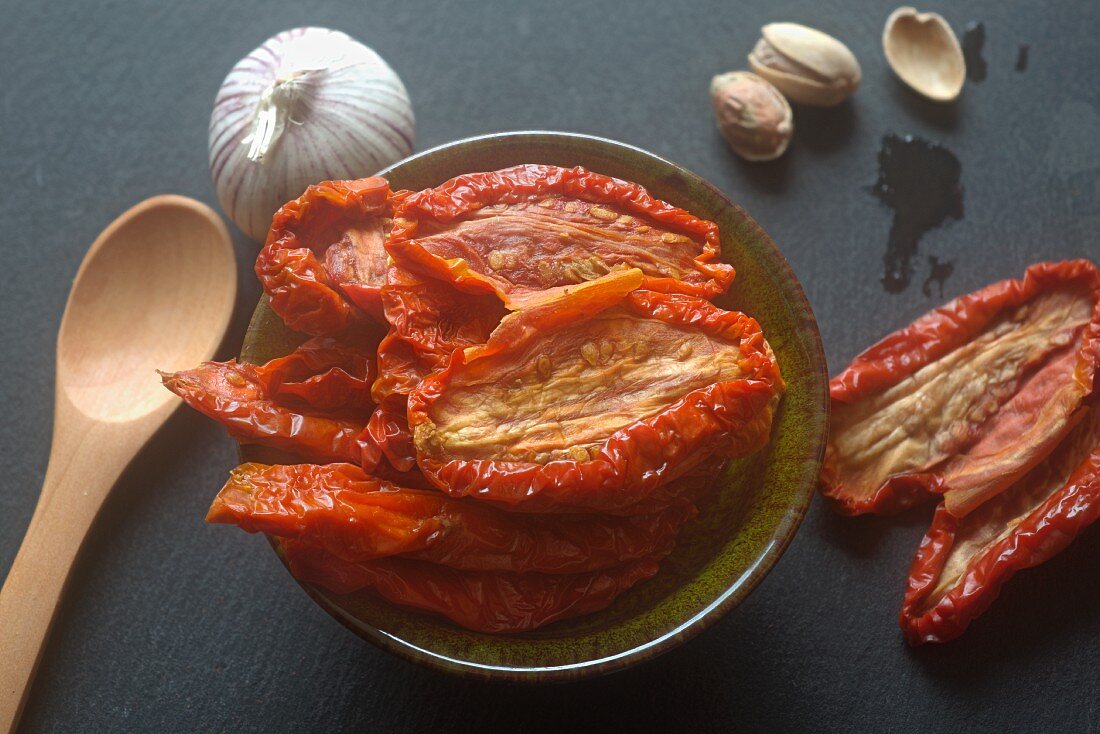 A bowl of dried tomatoes next to garlic and pistachio nuts