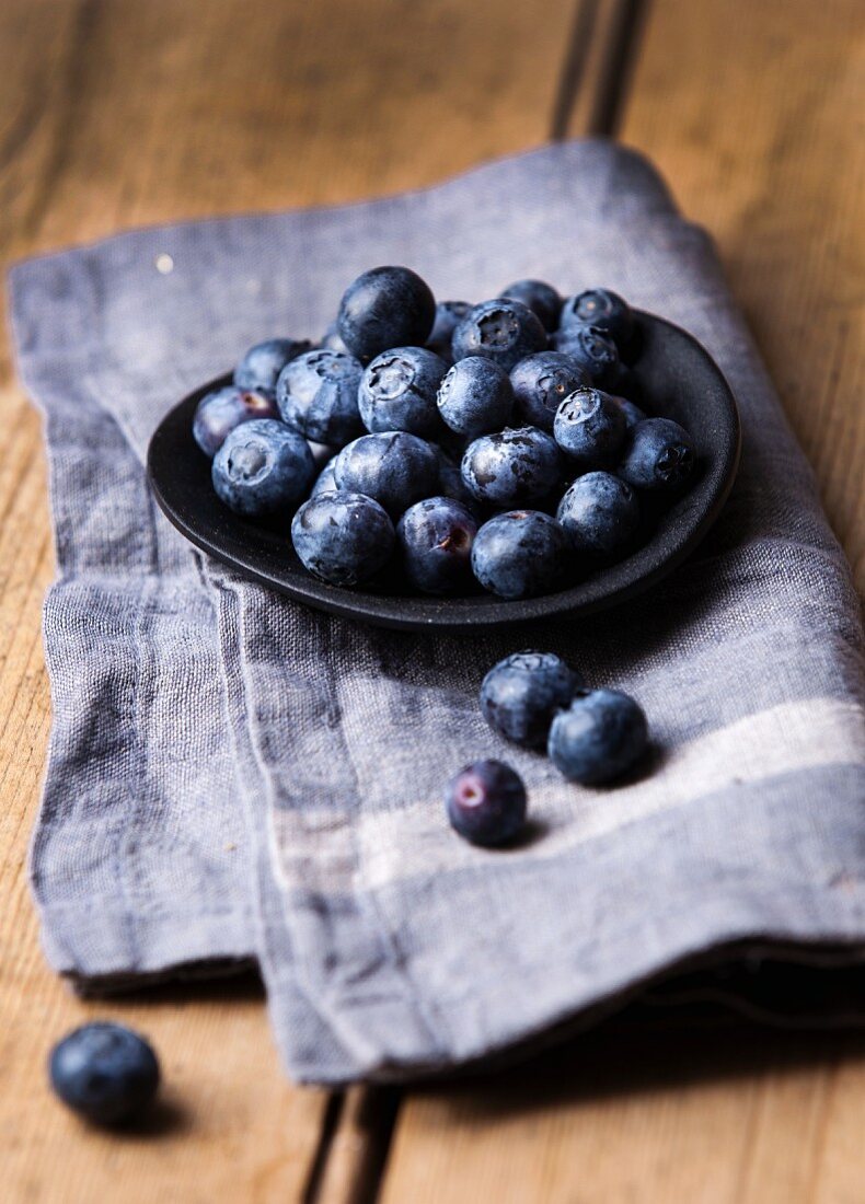 Heidelbeeren in einer flachen schwarzen Schüssel auf blauem Leinentuch