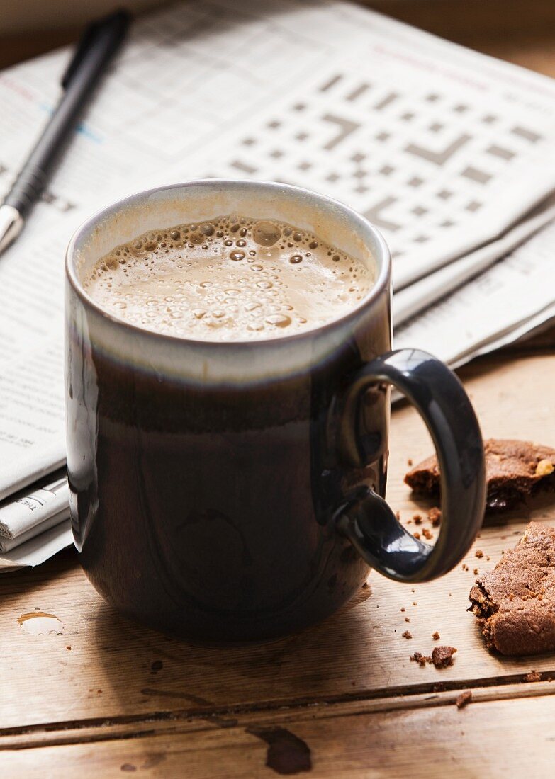 A cappuccino, a chocolate biscuit and a newspaper with a crossword on a wooden table