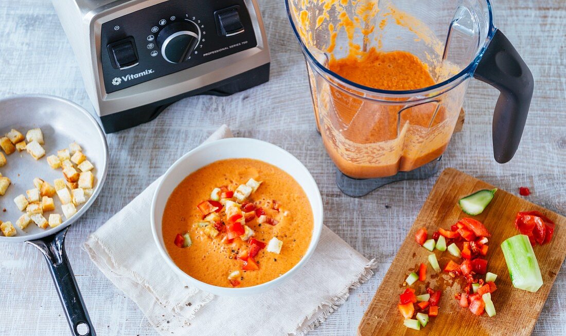 A bowl of gazpacho next to a blender, a pan and chopped vegetables