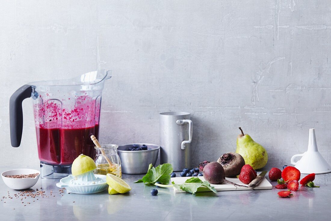 An arrangement of smoothie ingredients and a red smoothie in a blender