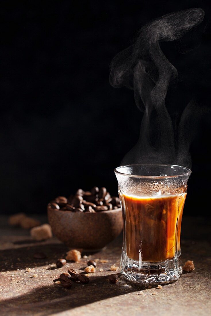 A steaming glass of coffee with coffee beans and sugar lumps in the background