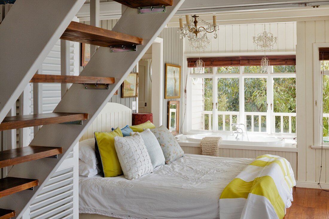 Double bed next to built-in bathtub below window in wood-panelled bedroom with staircase in foreground