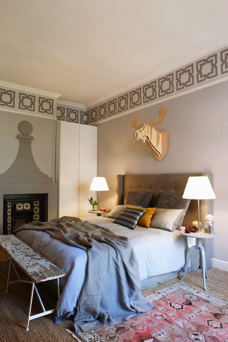 Double bed with upholstered headboard, table lamps on bedside tables, stencilled frieze on wall and stylised hunting trophy