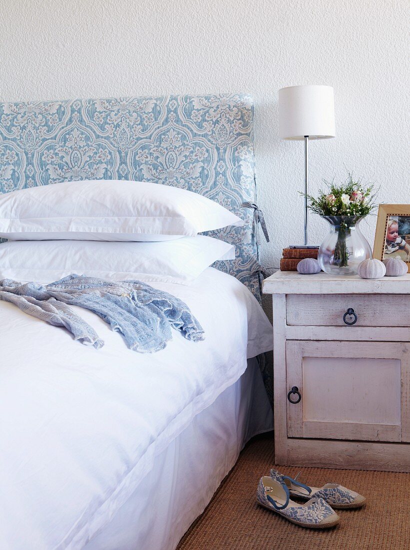 Bed with upholstered headboard and white bed linen next to bedside cabinet with table lamp and white lampshade; slippers on sisal carpet