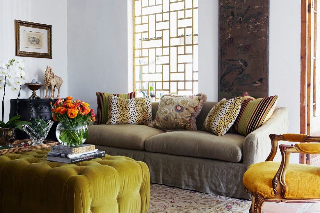 Ottoman with lime green cover and colourful bouquet in vase opposite sofa below window with wooden lattice framework; antique, upholstered armchair to one side