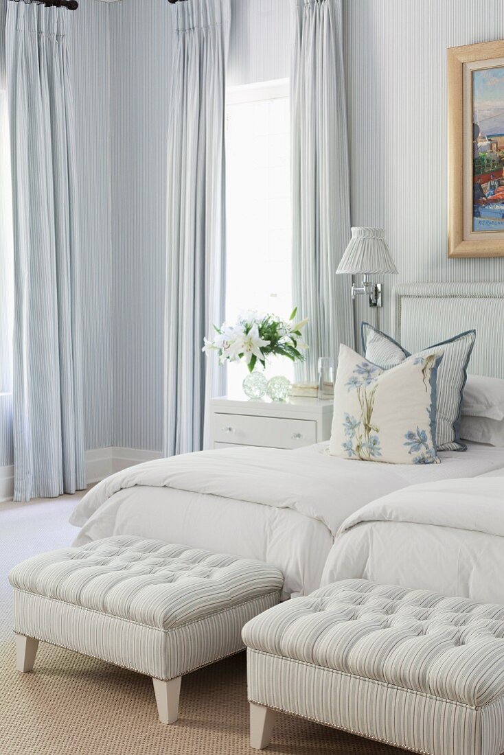 Ottomans with grey and white striped upholstery at feet of twin beds with headboards, curtains and wallpaper in the same pattern