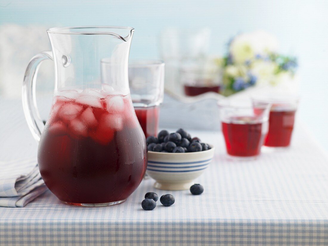 A jug of berry juice with ice cubes next to a bowl of blueberries