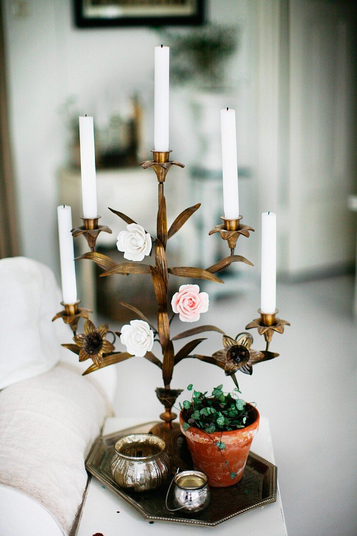 White candles in brass, floral candlestick with coloured rose ornaments on tray amongst potted plant and silver pots