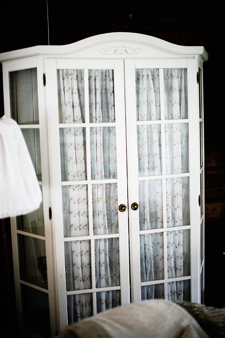 White-painted, glass-fronted cabinet with patterned curtains behind doors