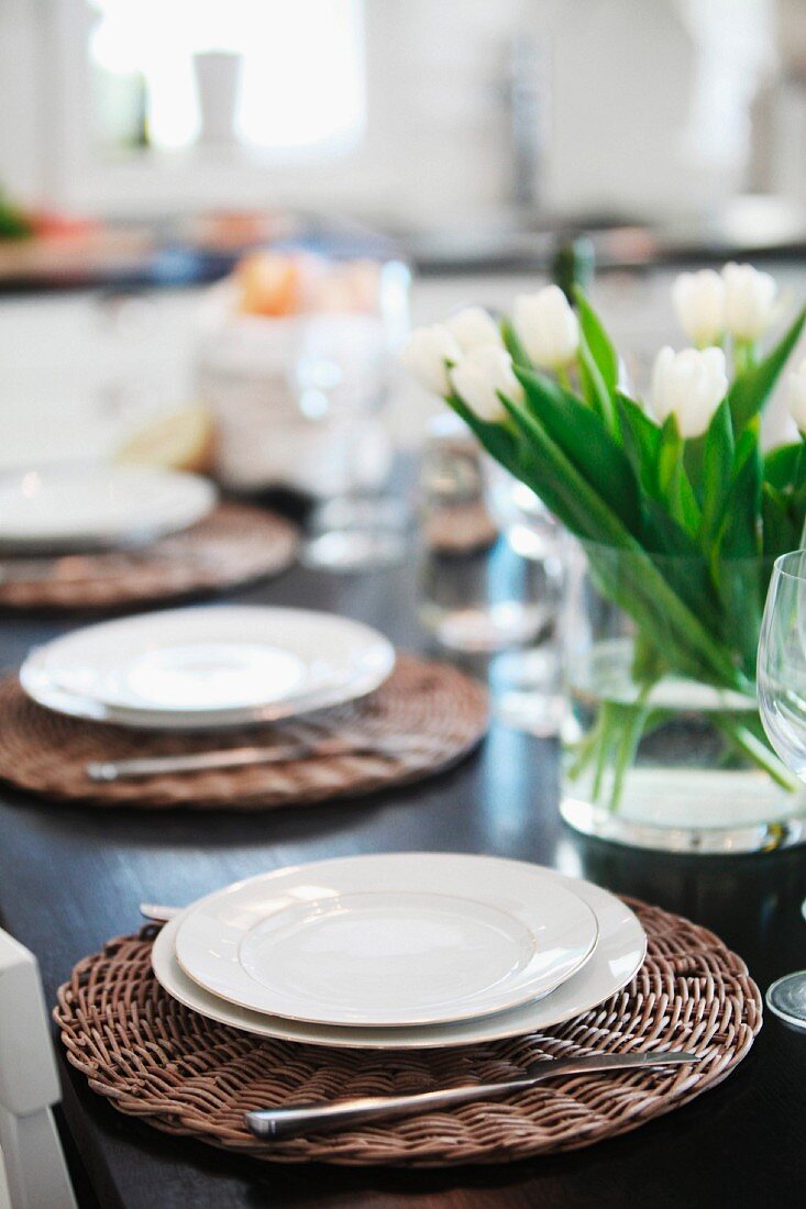 Table festively set with white plates on wicker place mats and glass vase of tulips
