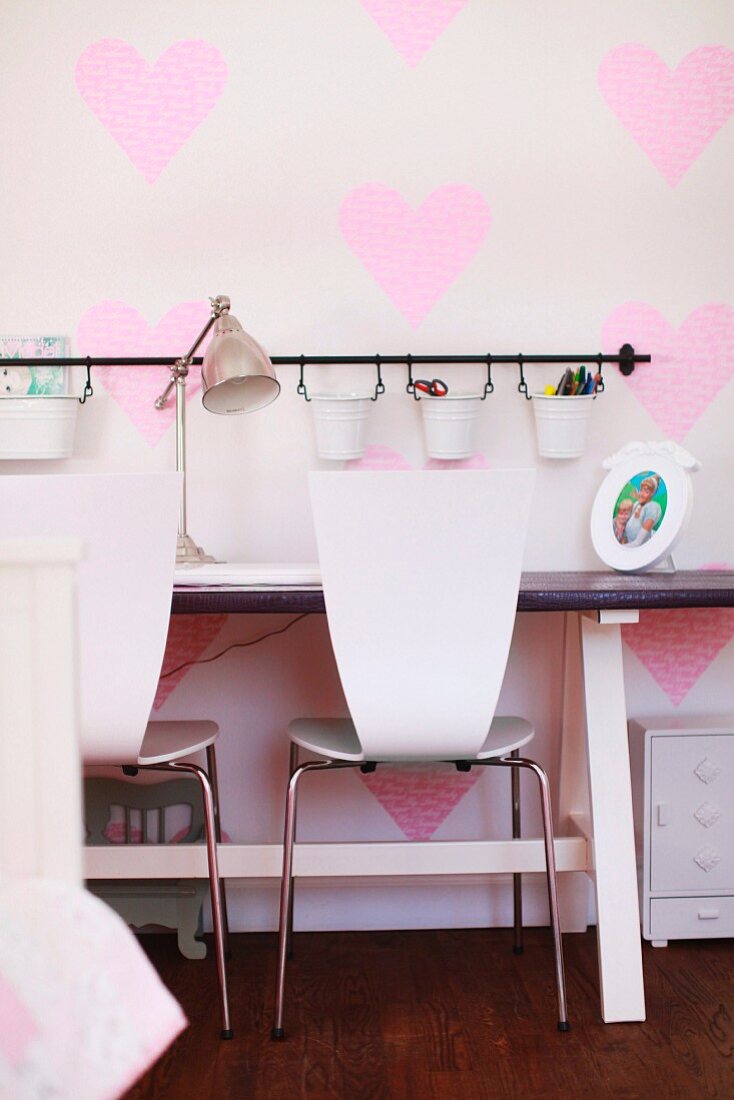 Scandinavian chairs at console desk with retro lamp; small metal buckets for bits and bobs hung on wall with heart-patterned wallpaper