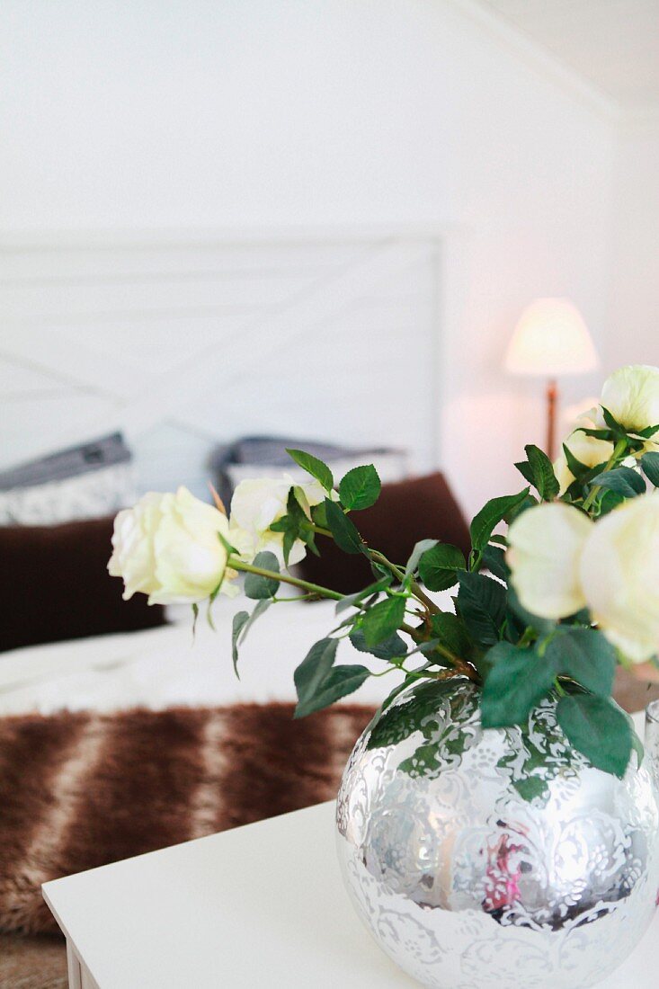 White roses in round, ornate, mercury glass vase; double bed in background
