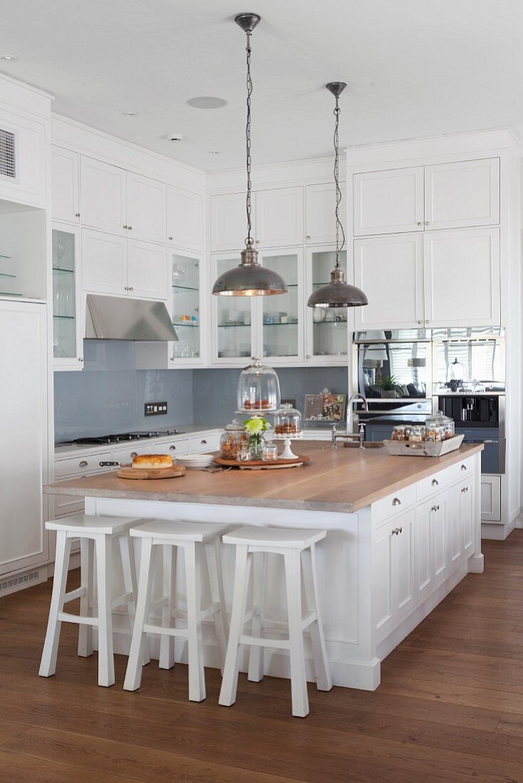 Free-standing island counter with white bar stools below pendant lamps with metal lampshades in open-plan country-house kitchen