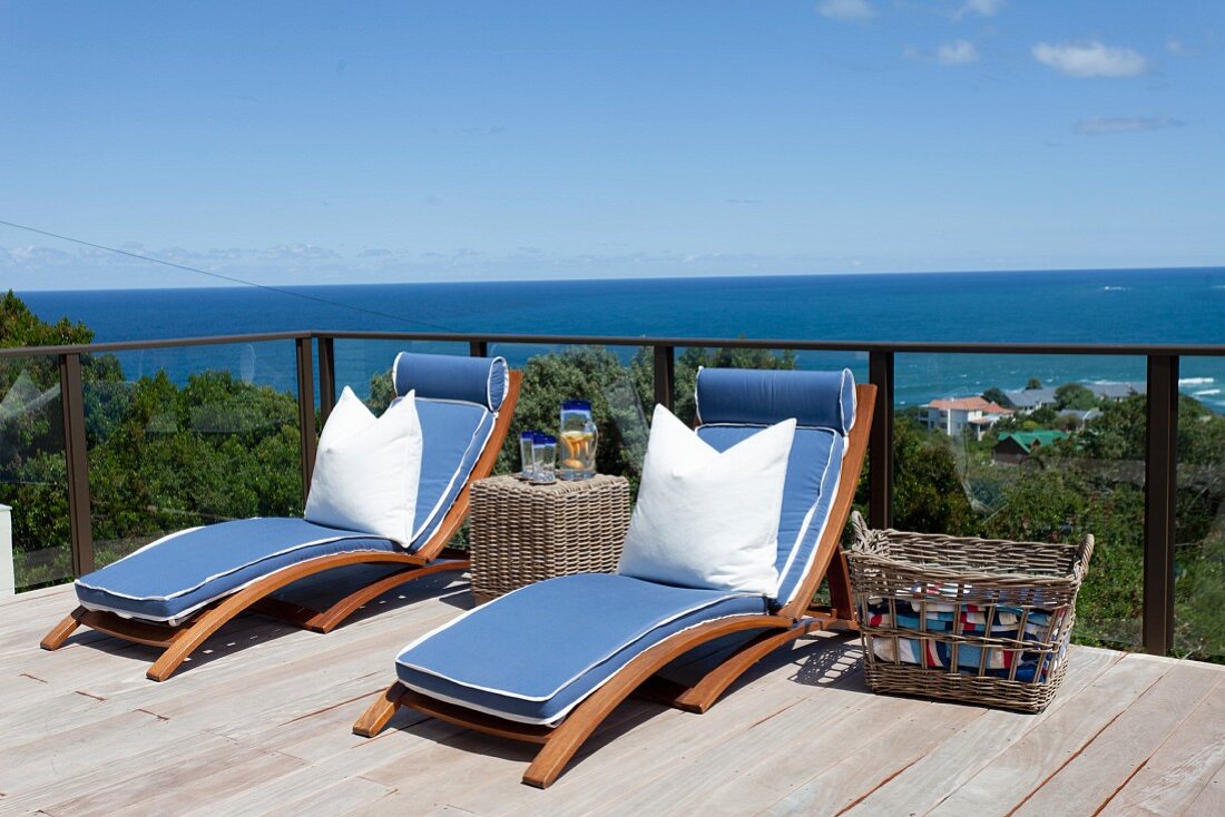 Sonnenliegen mit blauen Polstern und weißem Kissen auf Holzterrasse, über Geländer Panoramablick auf das Meer