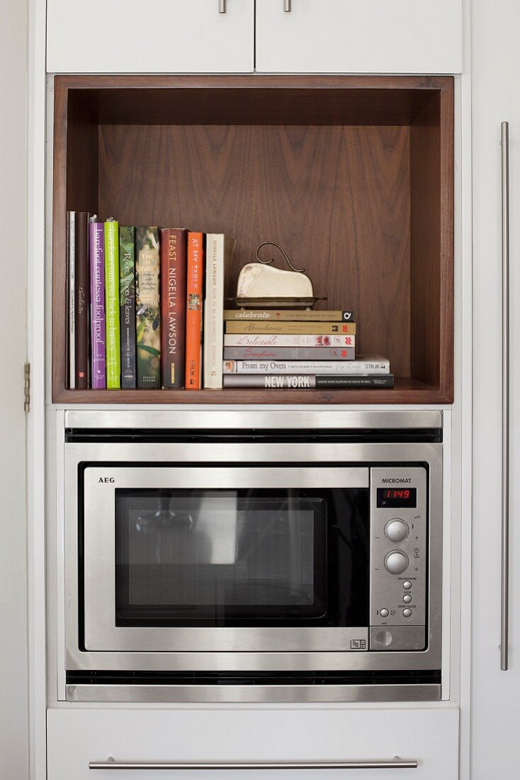 Microwave in kitchen cabinet below cookery books in wooden shelf compartment