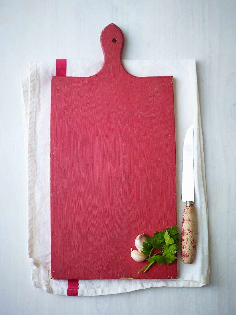 Rotes Schneidebrett aus Holz mit Knoblauch und Petersilie auf Geschirrtuch, daneben Messer