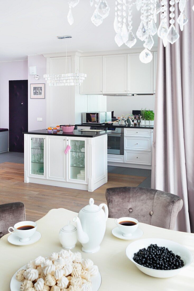 A view from a table laid for afternoon coffee looking towards an elegant, built-in white kitchen with an island counter and crystals from a chandelier at the top of the picture