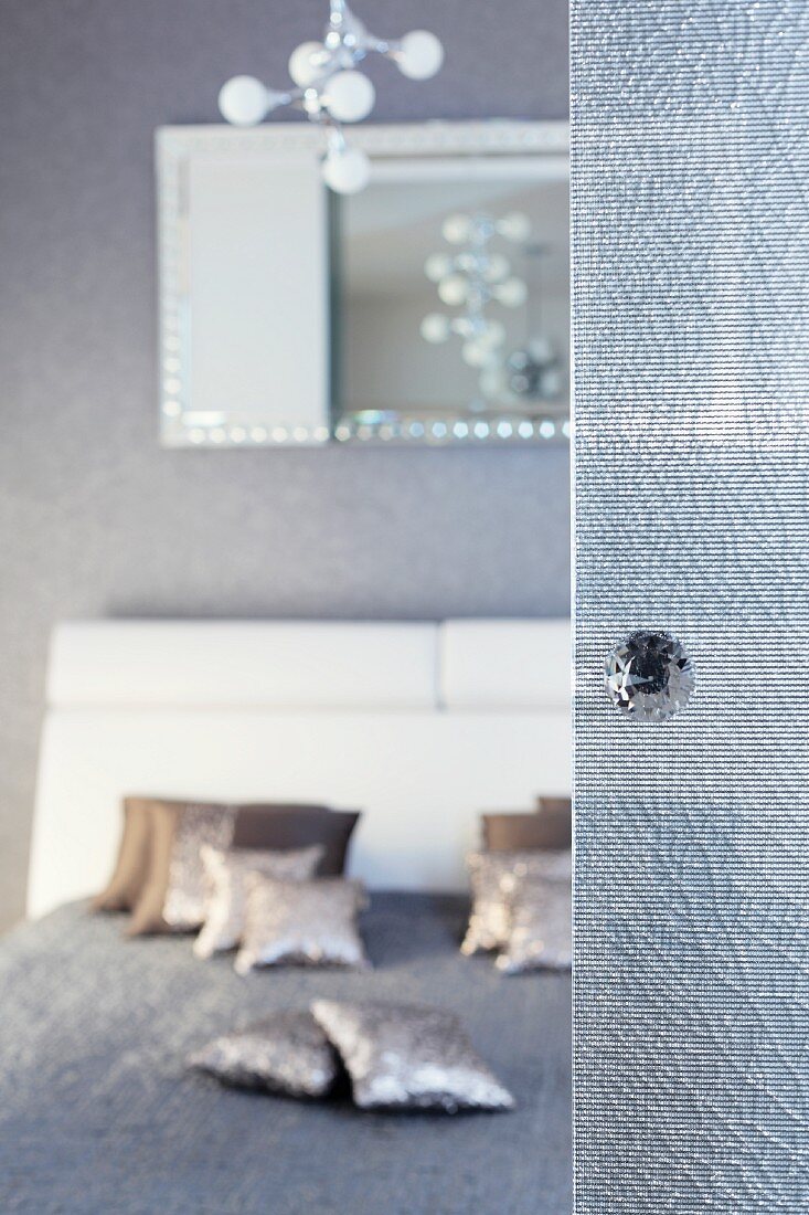 Glastür mit Knauf im Vordergrund; unscharfer Blick in den Schlafraum auf ein Doppelbett mit silbernen Dekokissen auf grauer Tagesdecke