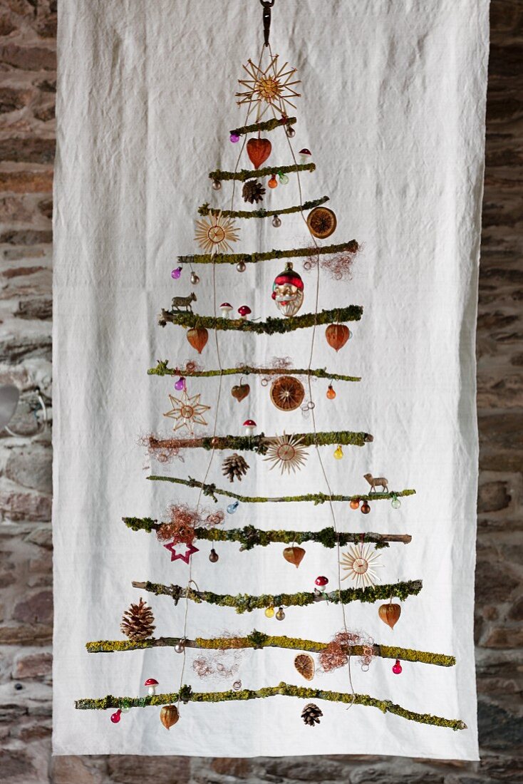 Stilisierter Weihnachtsbaum aus vermoosten Ästen mit weihnachtlichen Dekoartikeln vor weißem Leintuch