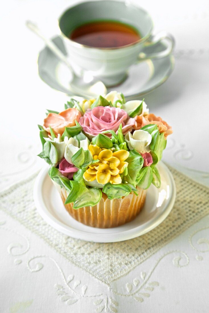 A cupcake decorated with romantic sugar flowers in front of a cup of tea