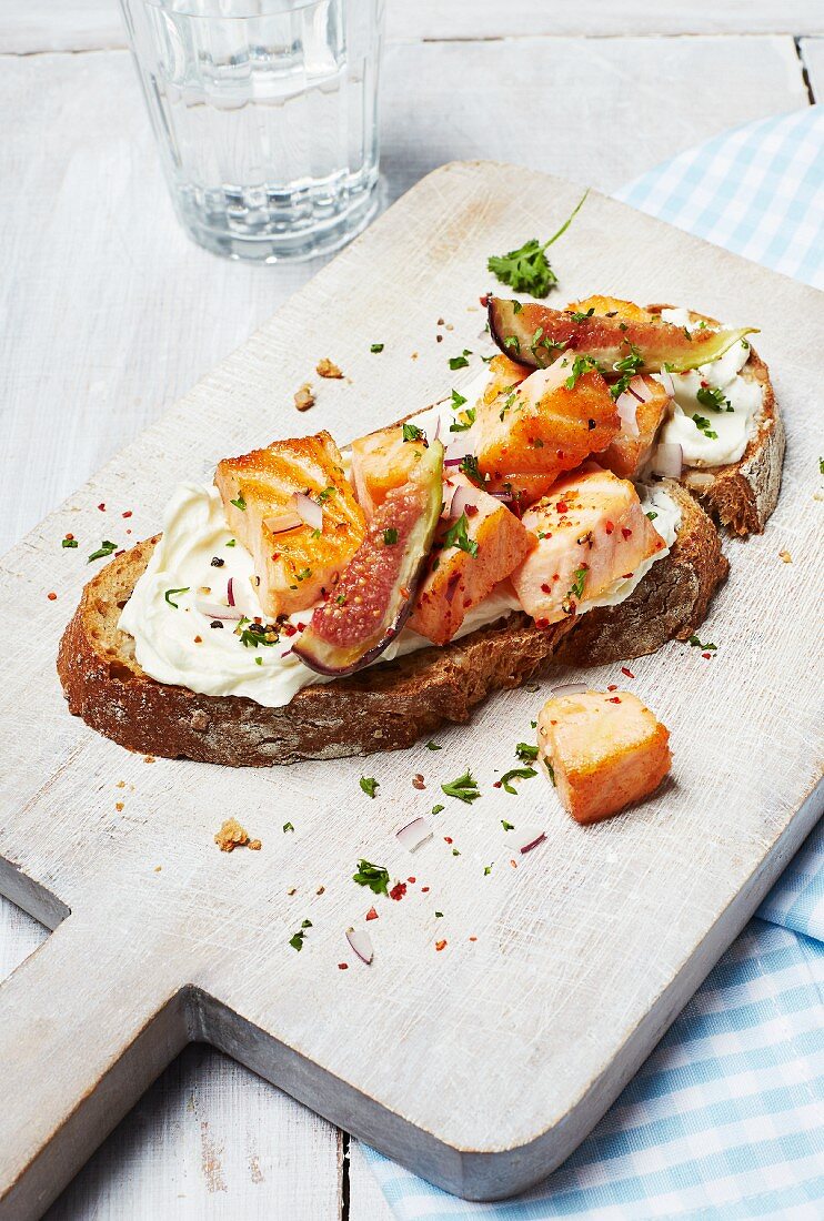 A slice of bread topped with salmon, cream cheese, figs and herbs on a chopping board