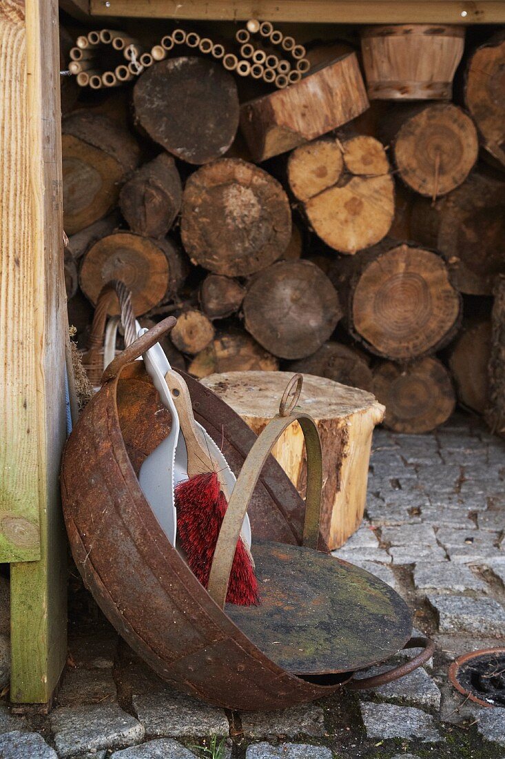 A dustpan and brush in a rusty bowl on cobbles in front of a wood shed