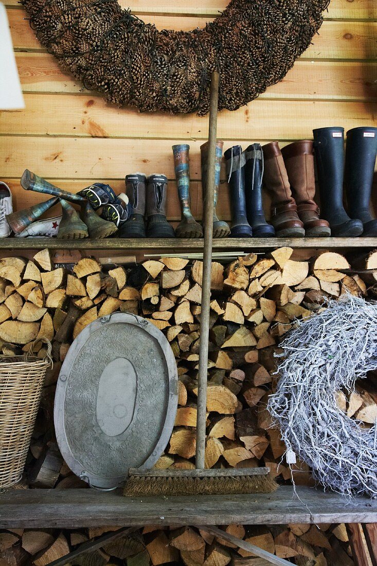 An old broom between a tray and a woven wreath on a wooden bench in front of a wood pile with a shelf of rubber boots above it