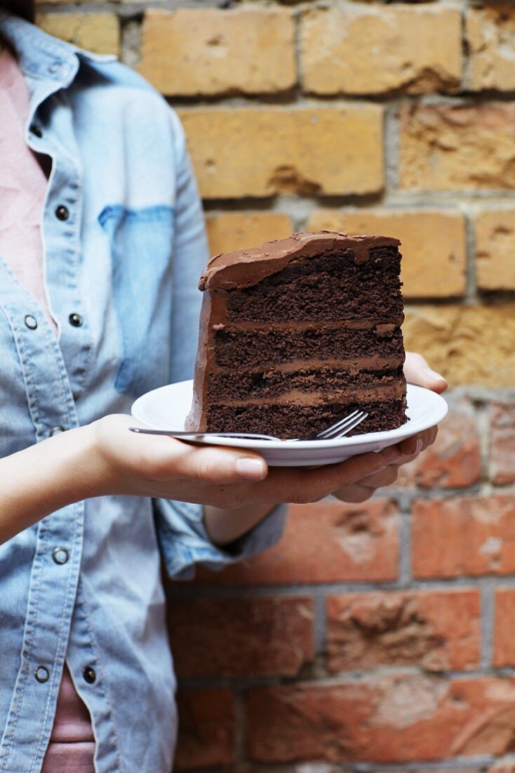 A person standing in front of a brick wall holding a large slice of chocolate cake on a plate