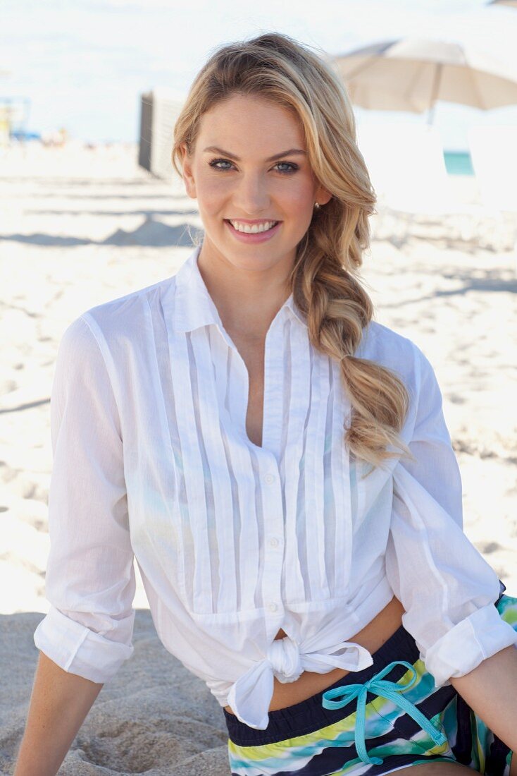 A young blonde woman wearing a white blouse and colourful shorts sitting on a beach