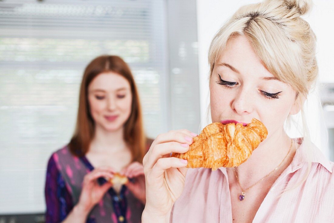 Two young women eating croissants in a kitchen