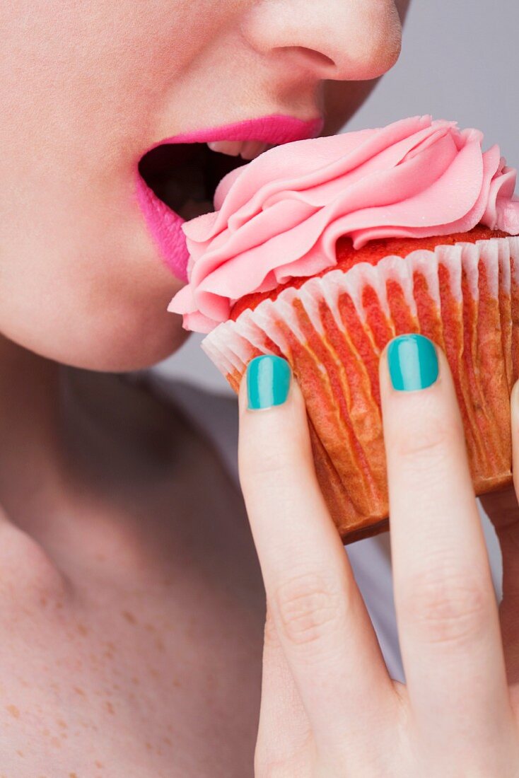 A young woman biting into a pink cupcake, face cropped