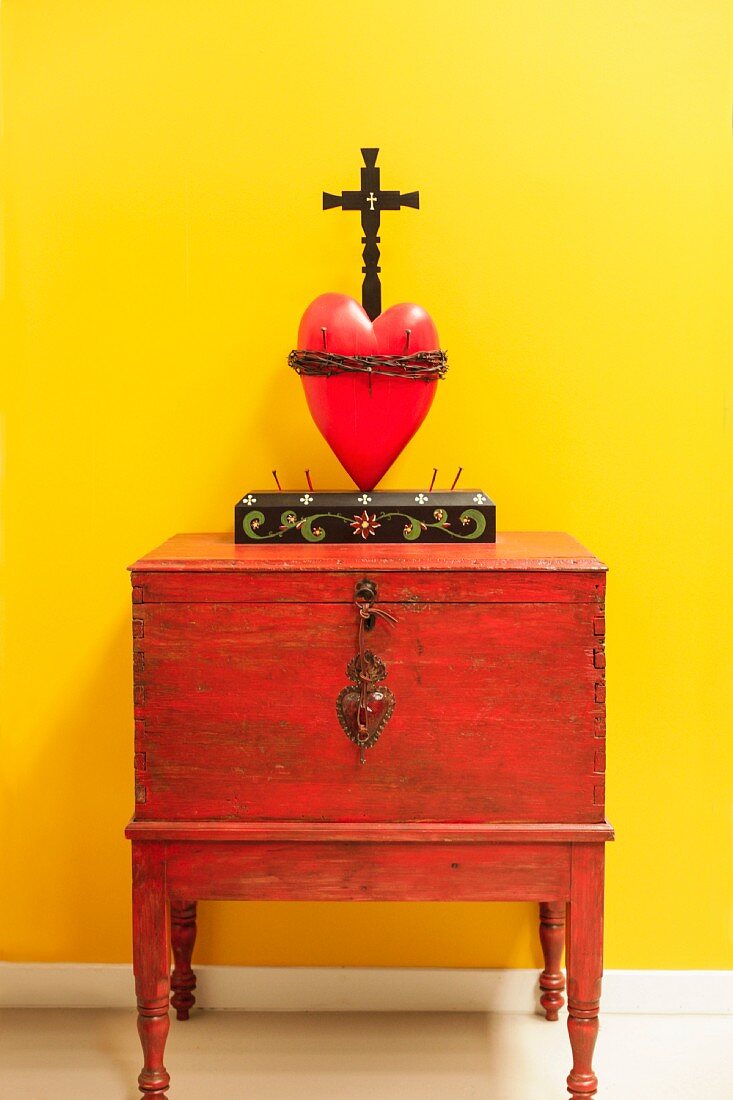 Kruzifix über rotem Herz auf Schränkchen vor leuchtend gelber Wand