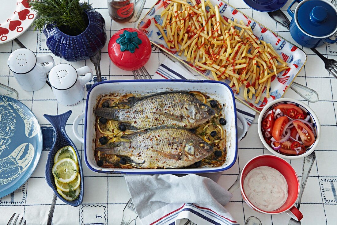 A view of a table with two roasted fish in a roasting tin, fries and salad