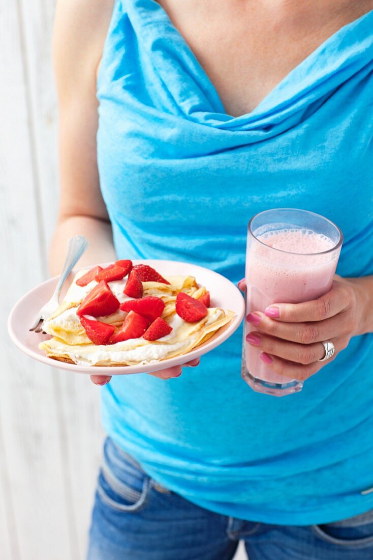 A woman holding a strawberry milkshake and pancakes with cottage cheese and strawberries