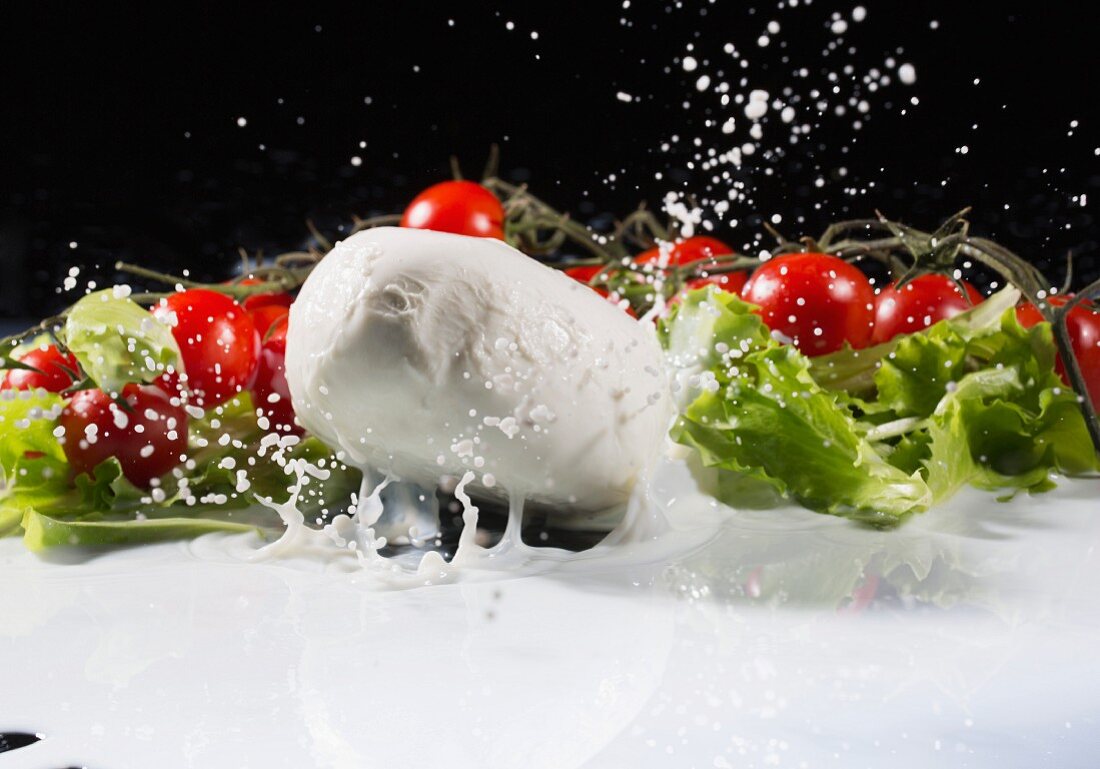 Mozzarella fällt spritzend in Milch, Strauchtomaten und Salatblätter im Hintergrund