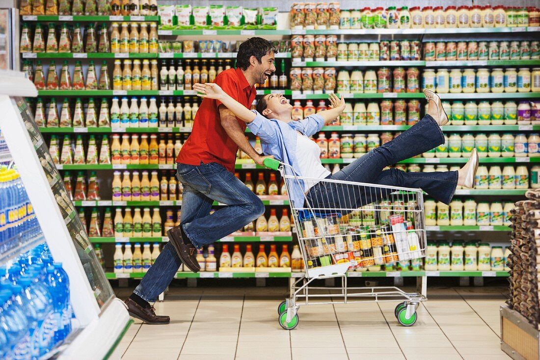 Mann schiebt Frau im Einkaufswagen durch Supermarkt