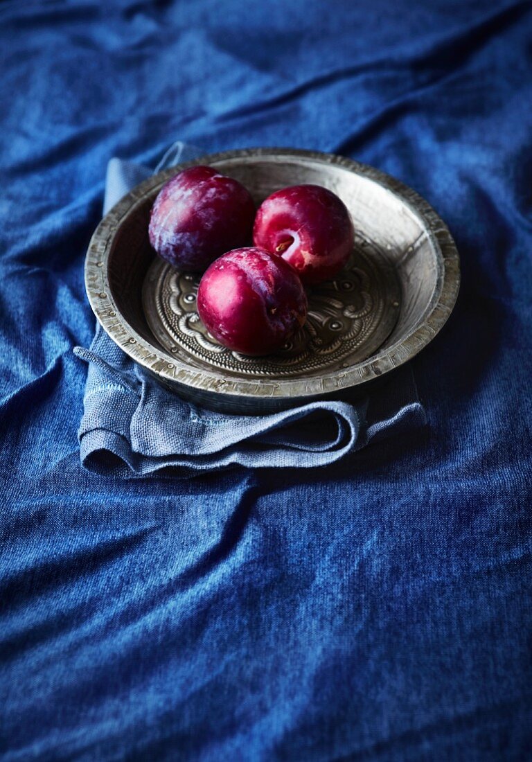 Three plums on a pewter plate on a blue cloth