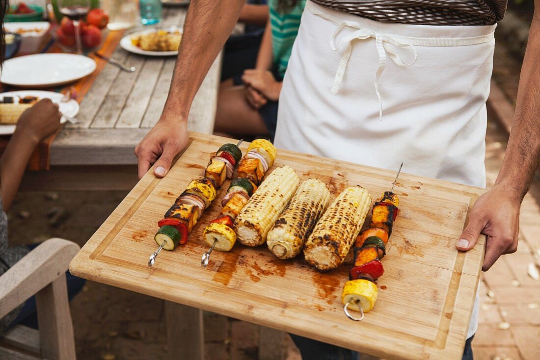 A man holding a wooden board of barbecued vegetable skewers and corn on the cob
