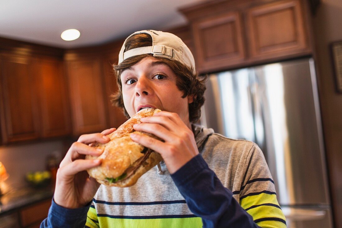 A teenage boy in kitchen eating a large sandwich