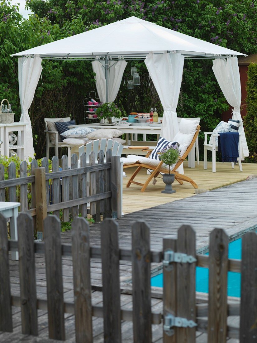 Zeltartiger Pavillon mit weissen, gerafften Vorhängen und Tisch-Bankgarnitur, in eingezäuntem Terrassenbereich