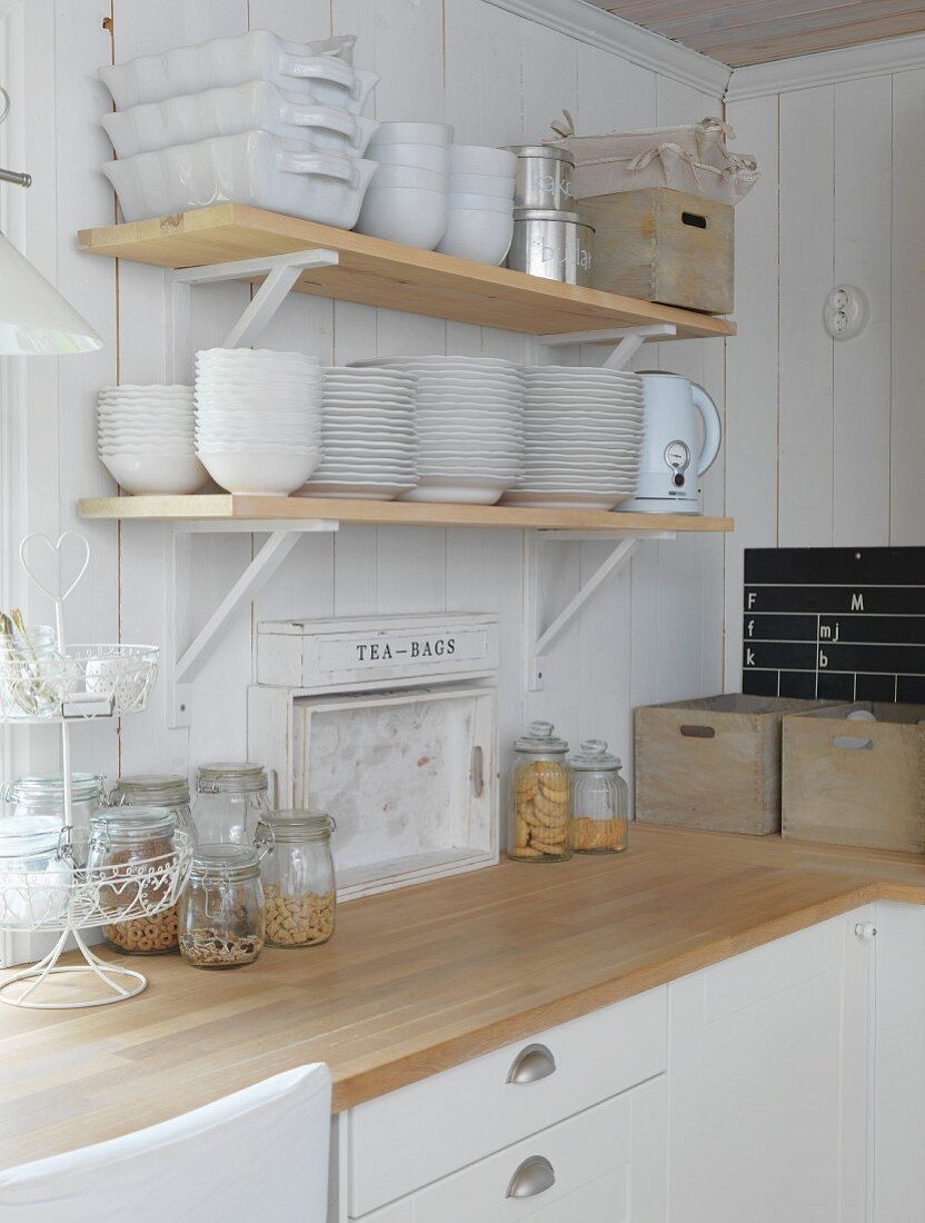 Corner of kitchen with crockery on bracket shelves on white wooden wall and storage jars on worksurface