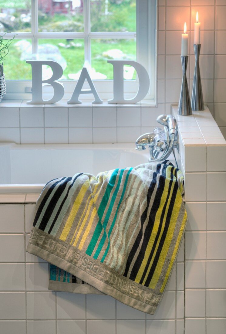 Striped towel, candles and decorative letters on white-tiled bathtub below window