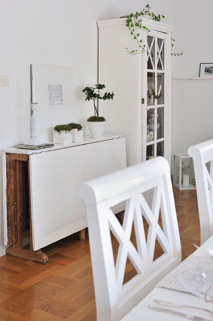 White wooden chairs at set table, ornaments on folding table used as console table and glass-fronted cabinet in background