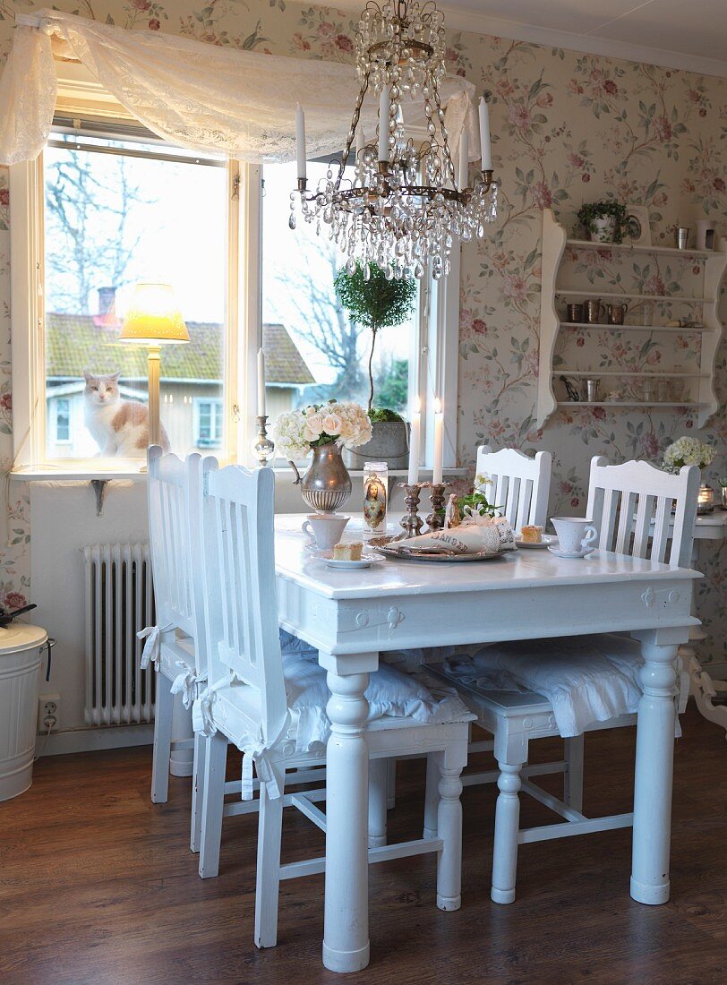 Dining area next to window; white-painted wooden table and chairs below chandelier with glass pendants