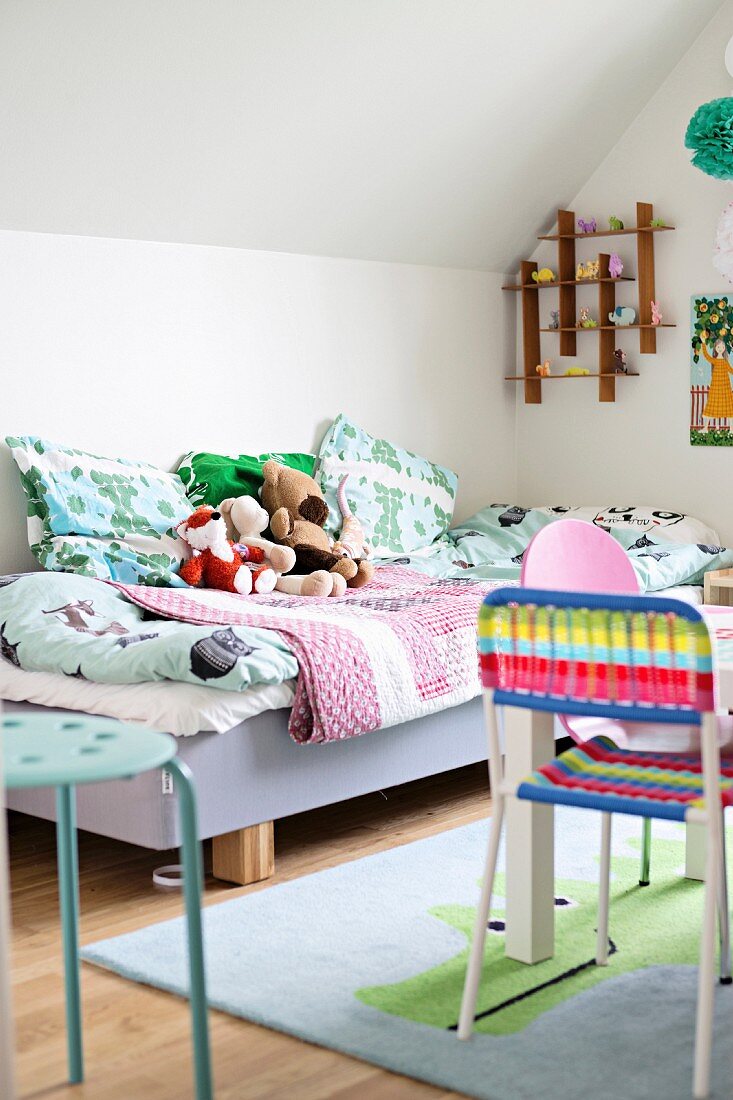 Kinderbett mit bunten Decken, Kissen und Kuscheltieren; bunter Stuhl an Spieltisch im Vordergrund